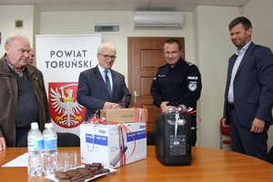 Starosta Toruński, Wicestarosta, Komendant Miejski Policji stoją w pokoju, a na stole leży sprzęt, który ma zostać przekazany Policji