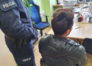 Policjant stoi przy zatrzymanym, który siedzi przy biurku podczas przesłuchania.