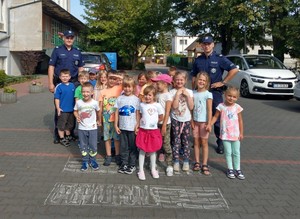 Policjanci z dziećmi przed placówką. Zdjęcie grupowe na namalowanym na placu przejściu dla pieszych.
