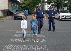 Dzieci przechodzą przez przygotowane przejście dla pieszych pod okiem policjantów.